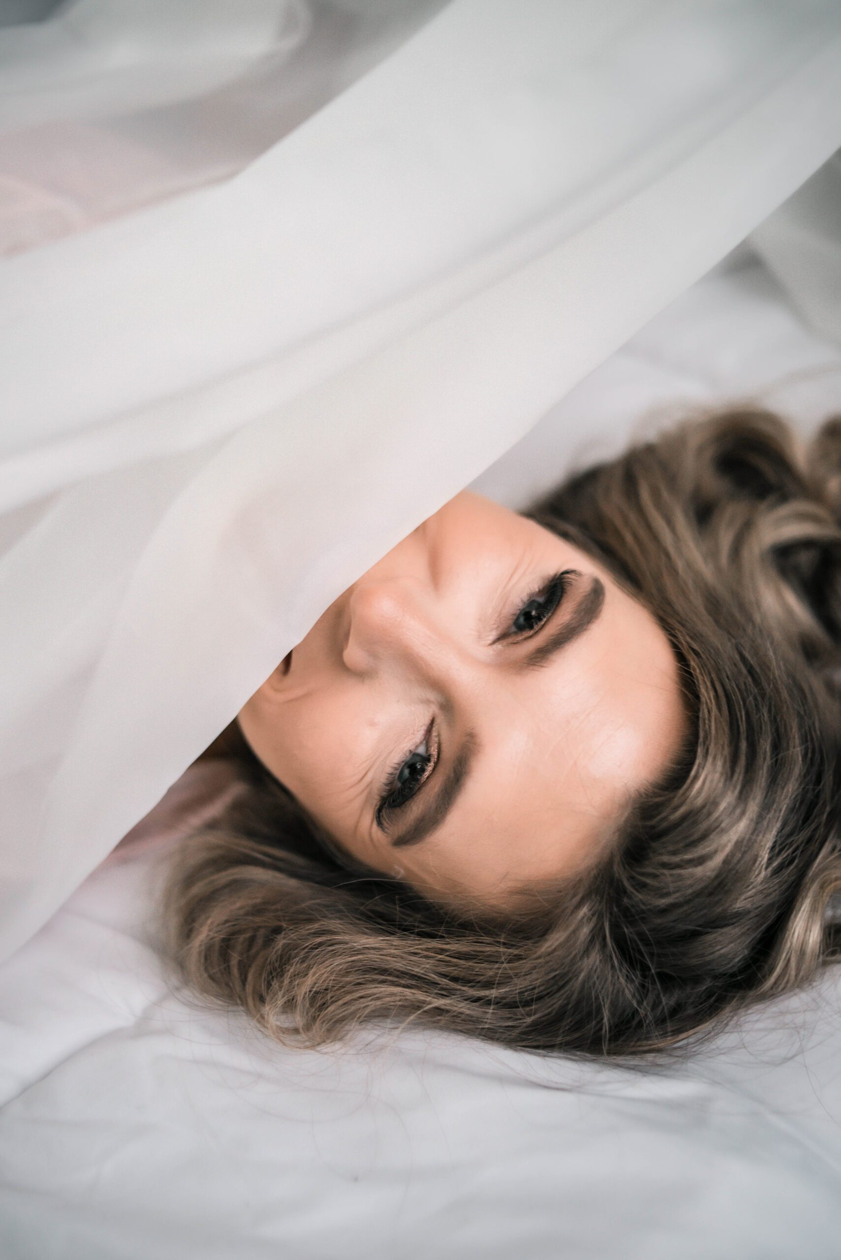 a smiling woman's face is covered by a sheer white fabric in a boudoir photoshoot