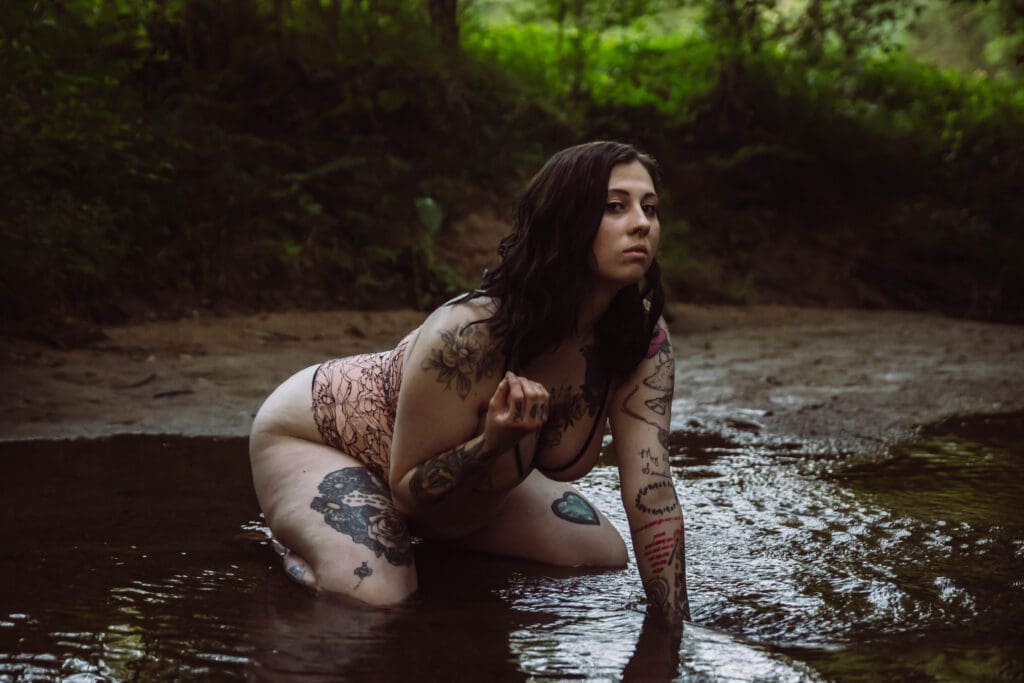 a woman with tattoos poses in a creek looking confident and daring